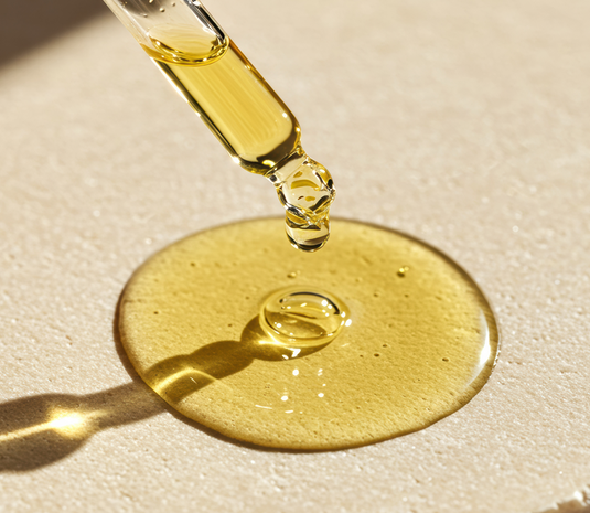 Gold dropper bottle with oil dripping onto a beige surface